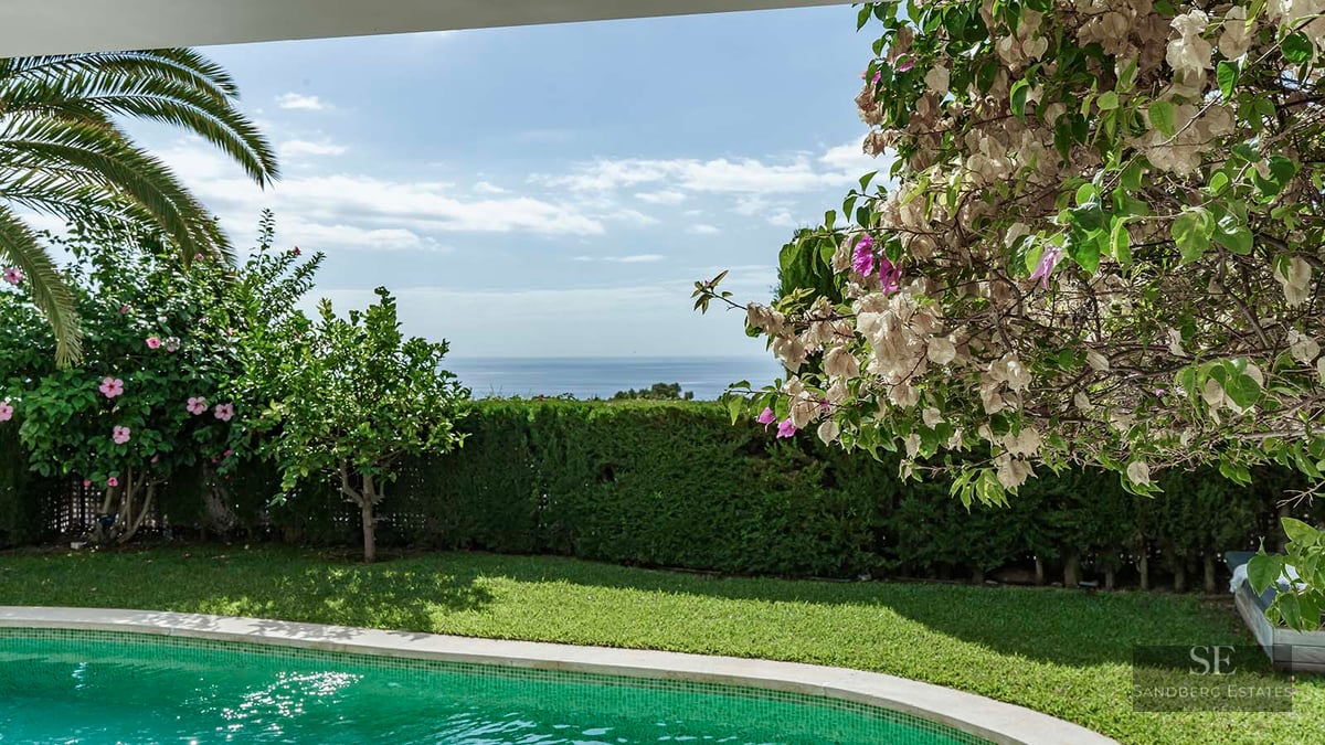 Turquoise swimming pool surrounded by green lawn and blooming flowers with panoramic sea views.