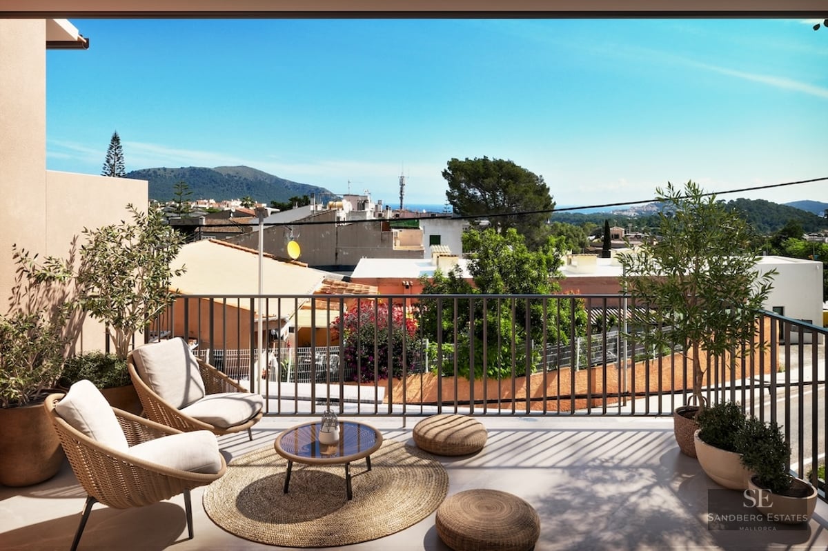 Sun-drenched terrace with stylish wicker chairs, potted olive trees, and views over a coastal town toward the mountains.