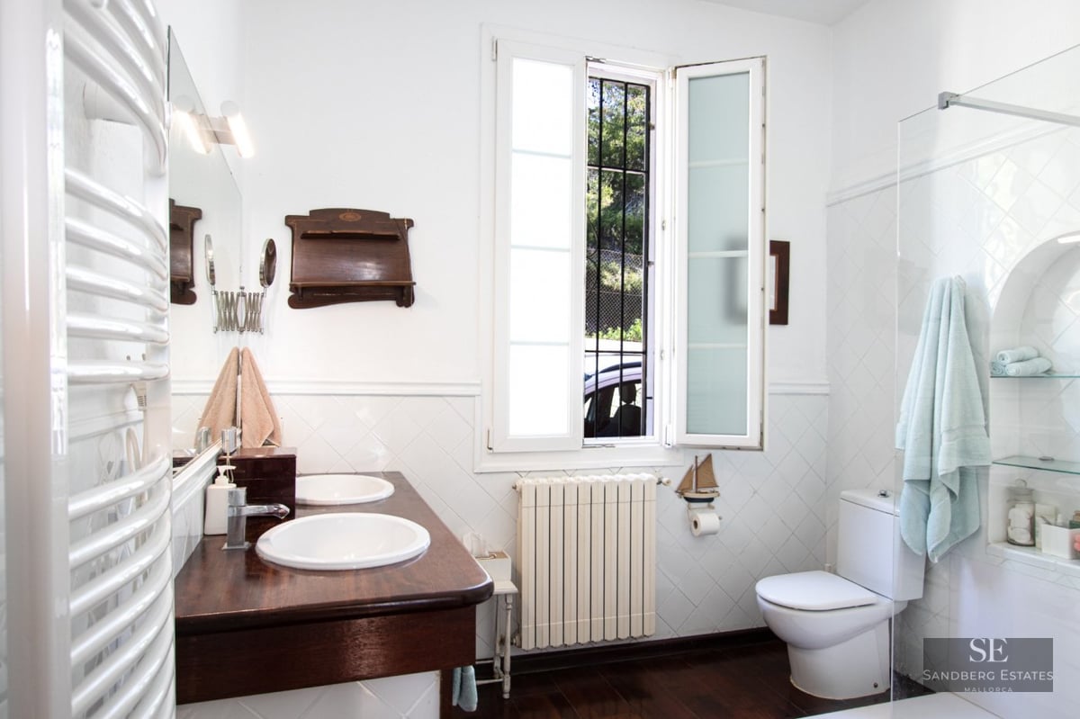 A bathroom featuring a double white sink vanity on dark wood, white tiled walls, a window, and a white toilet.