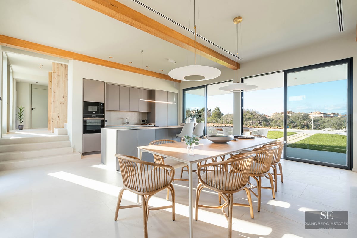 Bright dining room with wooden table and chairs next to a minimalist kitchen with large glass doors to garden.