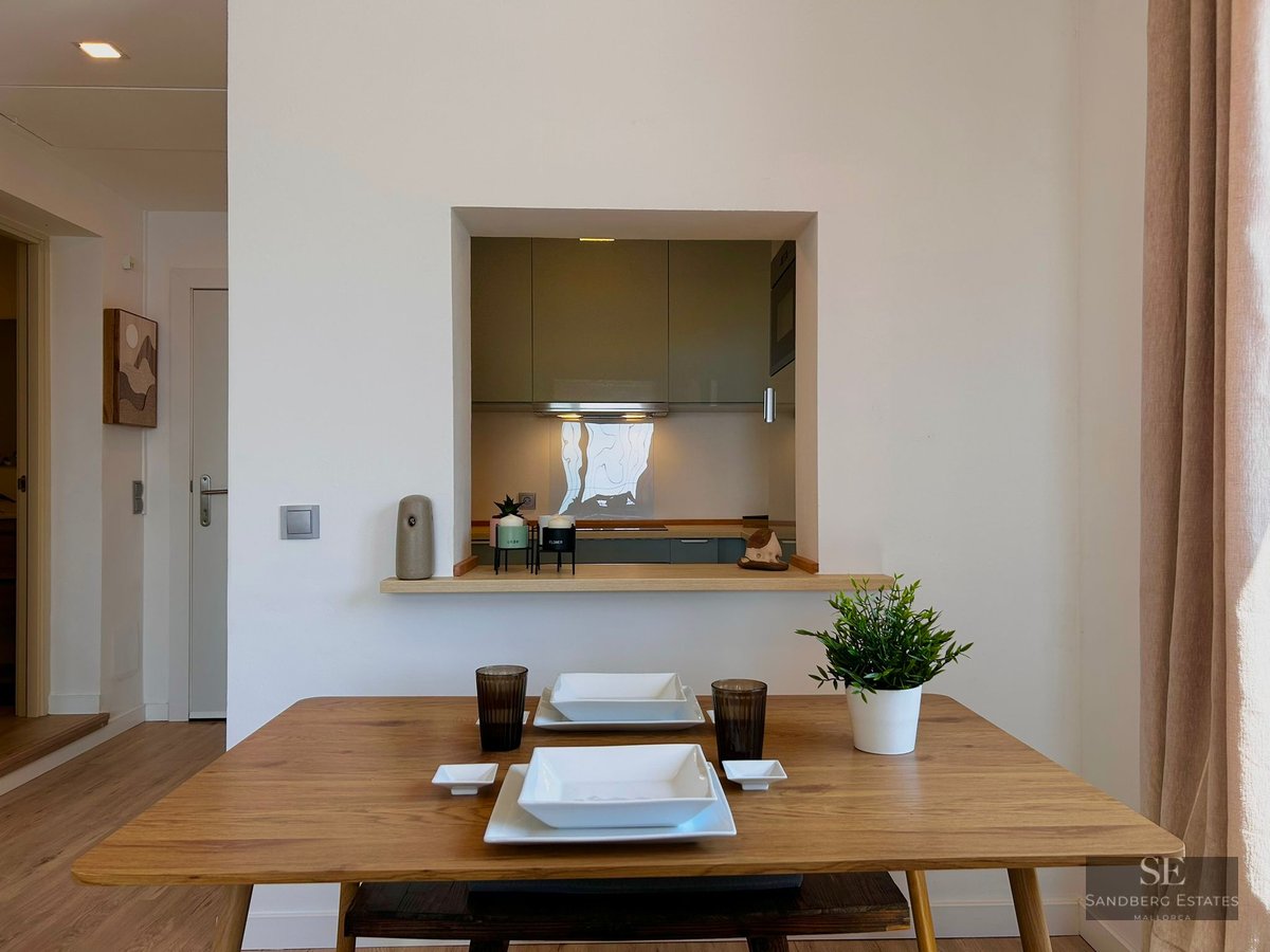 Wooden dining table set for two in front of a wall opening looking into a modern kitchen.