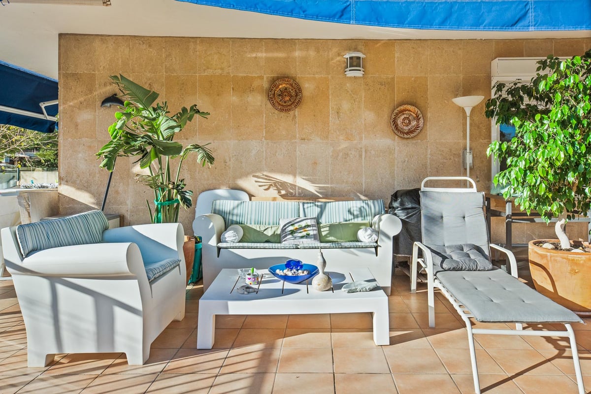 Sonnige überdachte Terrasse mit weißen Loungemöbeln, einer strukturierten Steinwand und üppigen Topfpflanzen.