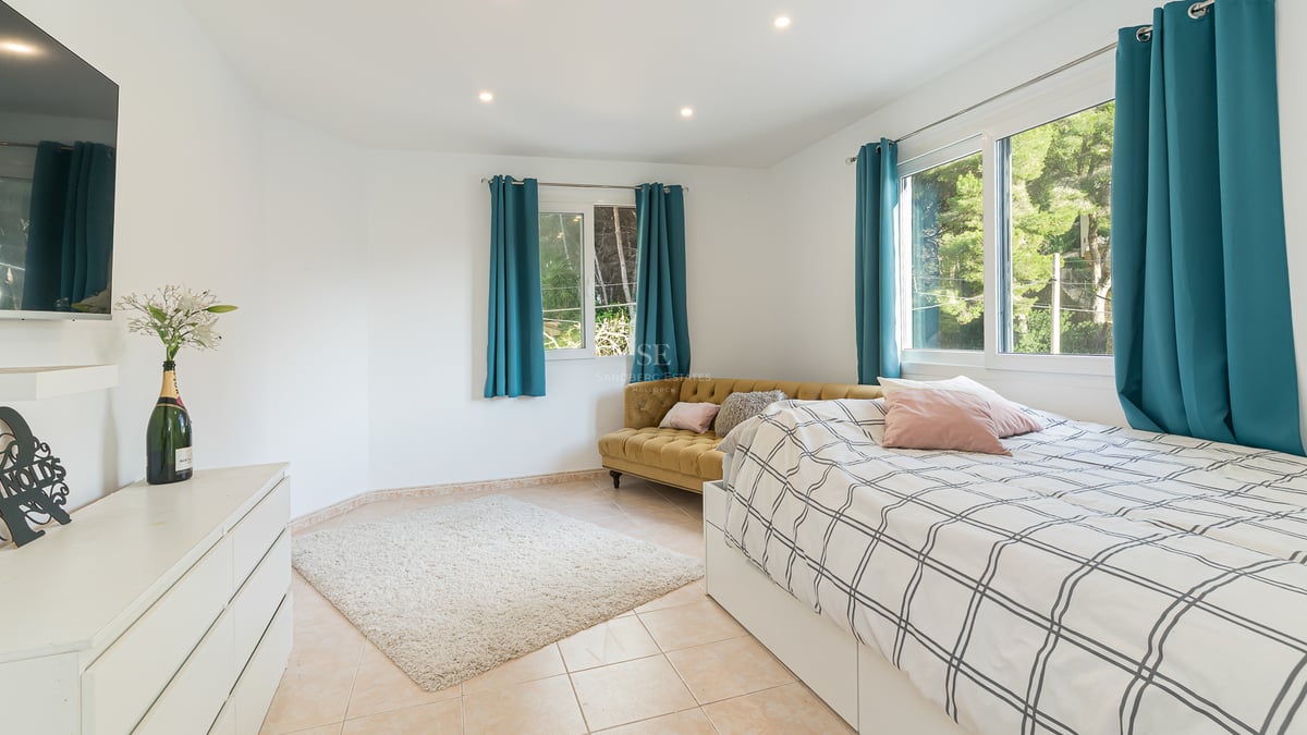 Chambre lumineuse avec lit, canapé en velours moutarde, rideaux bleu canard et vue sur la forêt par les fenêtres.