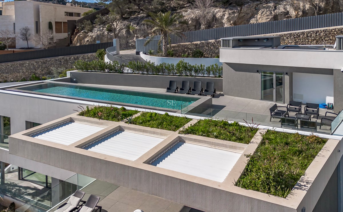 Vue aérienne d'une villa moderne grise avec piscine à débordement, chaises longues et toit végétalisé devant une colline.