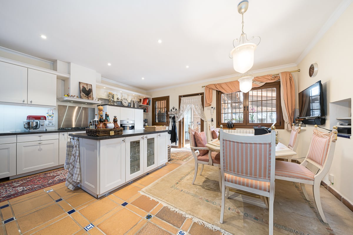 Grande cuisine avec meubles blancs, îlot central, sol en terre cuite et coin repas avec chaises rayées.