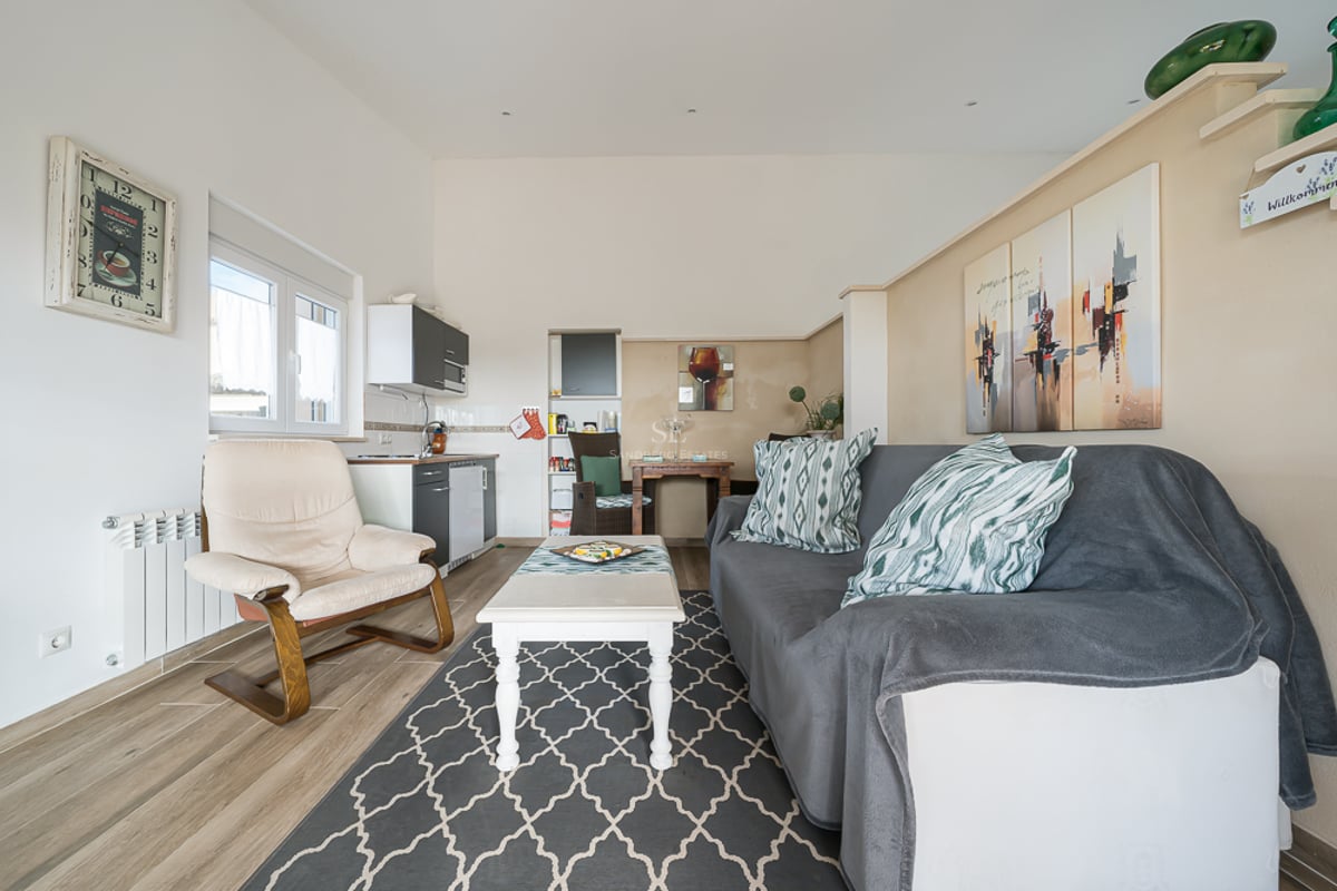 Salon lumineux avec canapé gris, fauteuil crème, tapis à motifs et kitchenette moderne sur murs blancs.