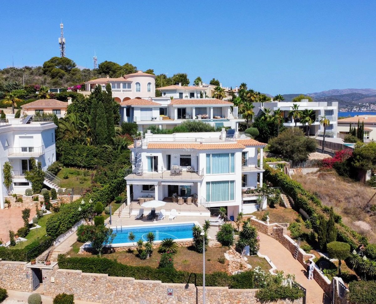 Vue aérienne d'une villa blanche à plusieurs niveaux avec une piscine turquoise, des murs en pierre et des jardins.