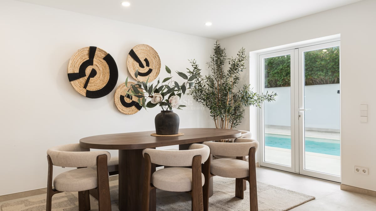 Oval wooden dining table with beige chairs in front of glass doors showing a swimming pool.