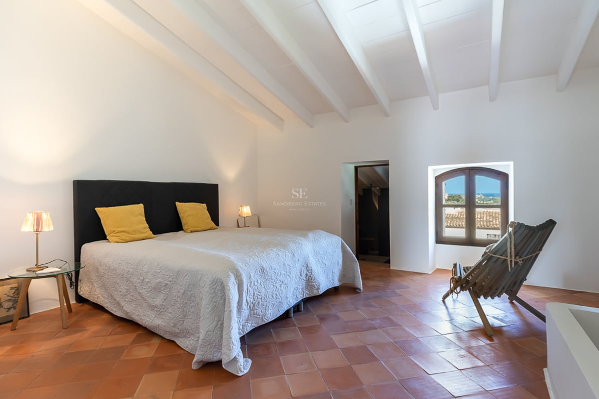 Chambre spacieuse avec poutres blanches, sol en terre cuite et fenêtre cintrée donnant sur la mer.