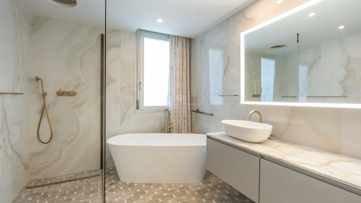 Luxury bathroom featuring marble walls, a freestanding white tub, brushed gold fixtures, and a large backlit mirror.
