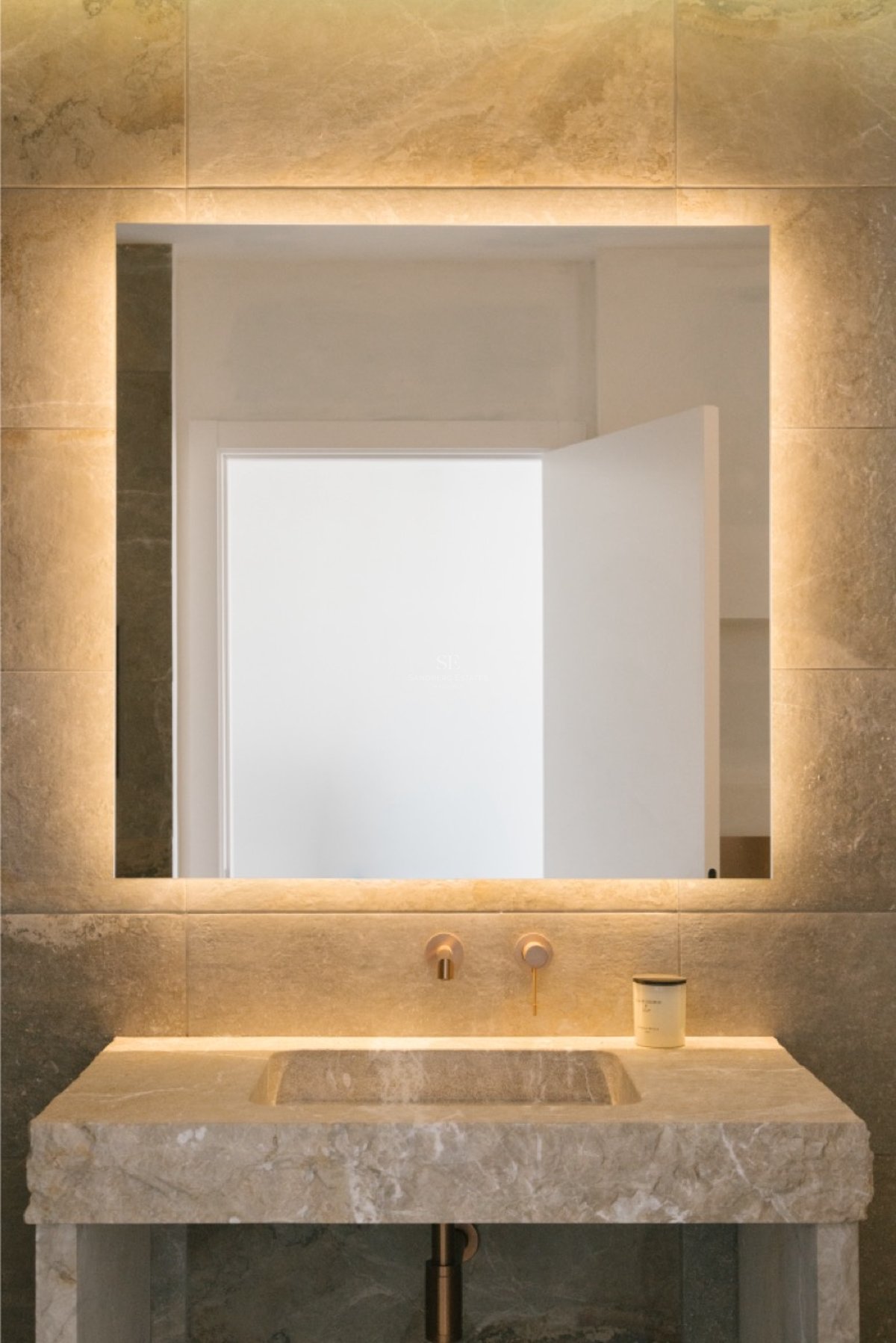 Nahaufnahme eines Waschtischs aus Naturstein mit integriertem Becken, Wandarmaturen aus Bronze und LED-Spiegel.