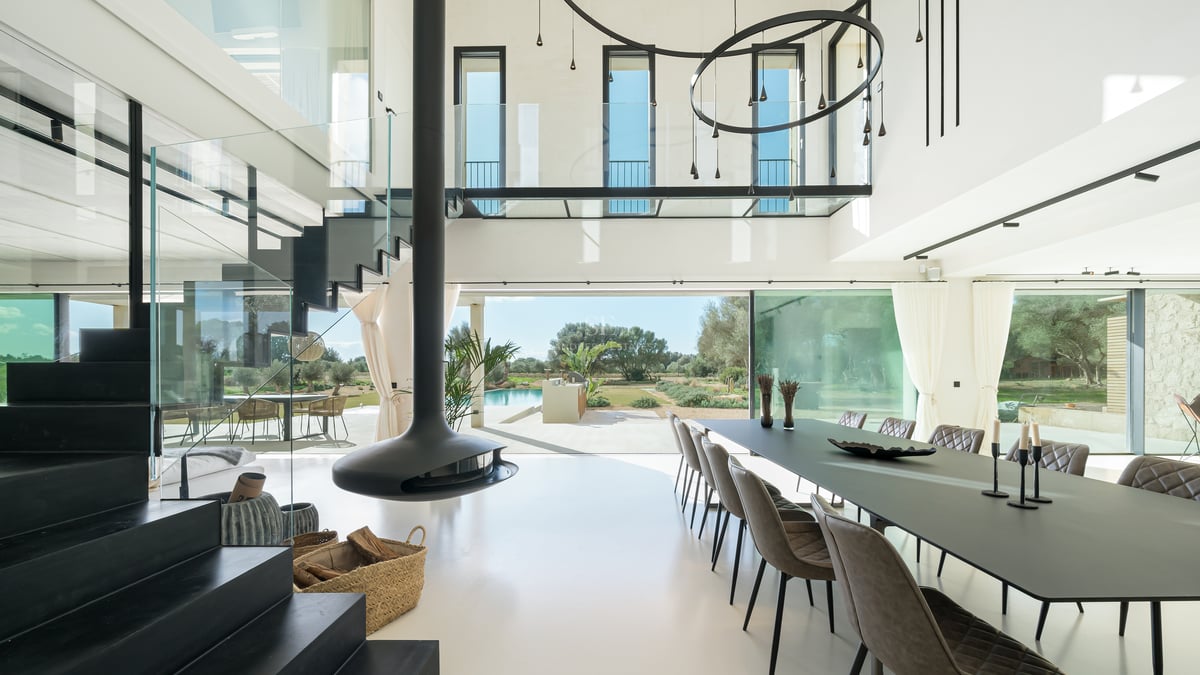 Ljus matsal med dubbel takhöjd, svart hängande öppen spis, glasräcken och utsikt över trädgård och pool.