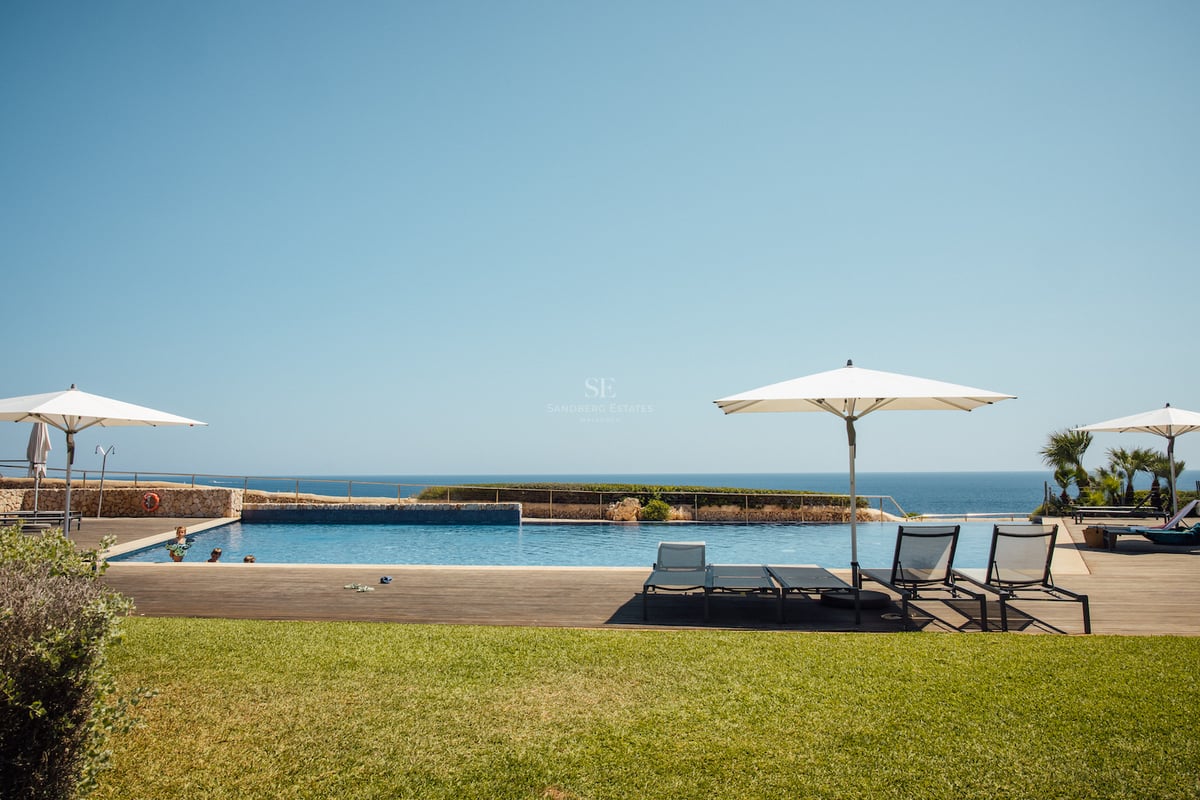 Grande piscine extérieure avec terrasse en bois et chaises longues face à la mer sous un ciel bleu.