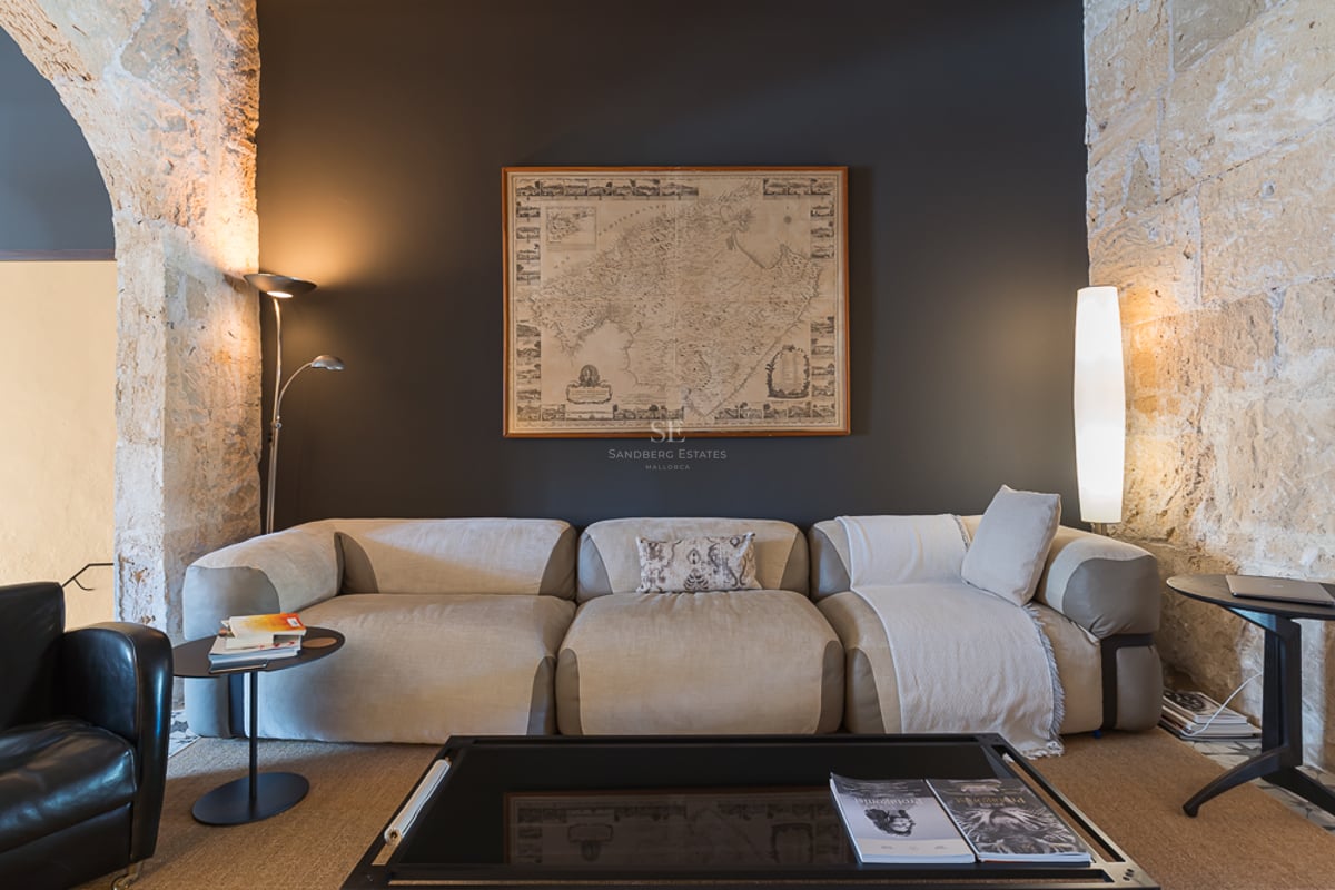 Canapé beige moderne devant un mur anthracite avec carte de Majorque et arches en pierre apparente.