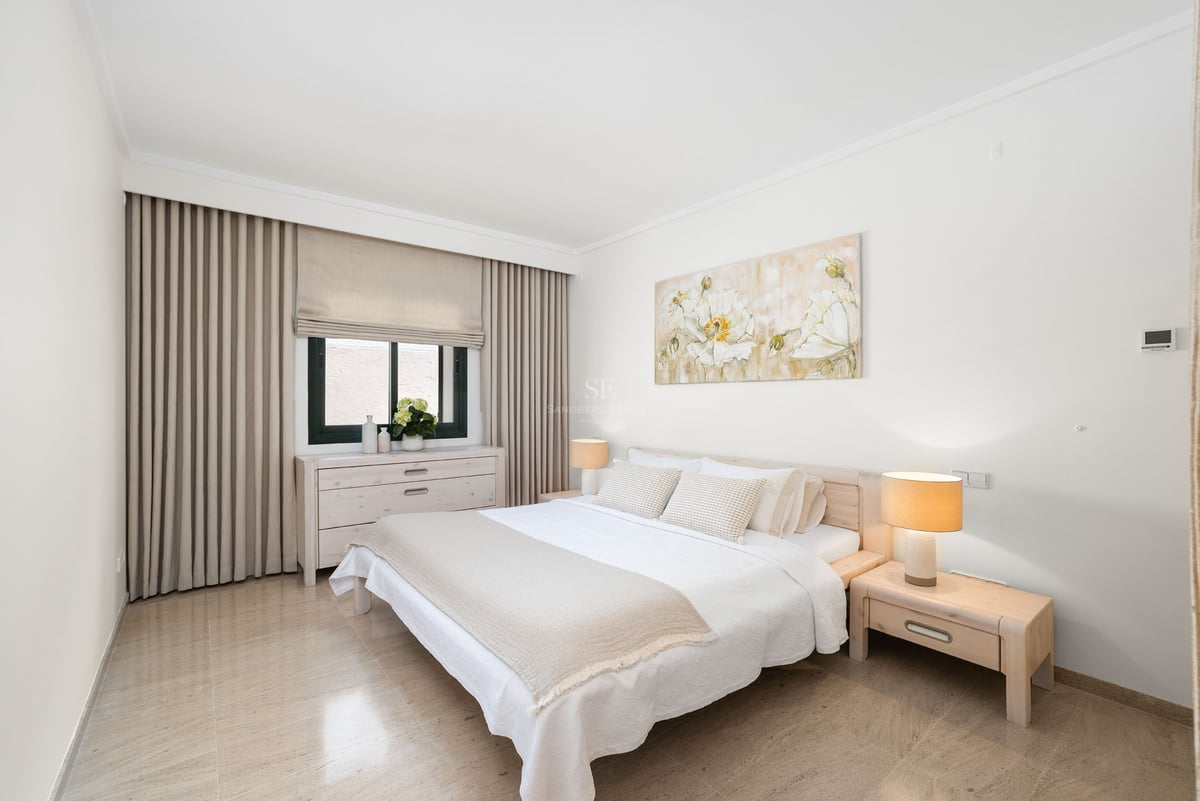 Chambre lumineuse avec lit blanc, tables de chevet en bois, tableau floral et sol en carrelage beige.
