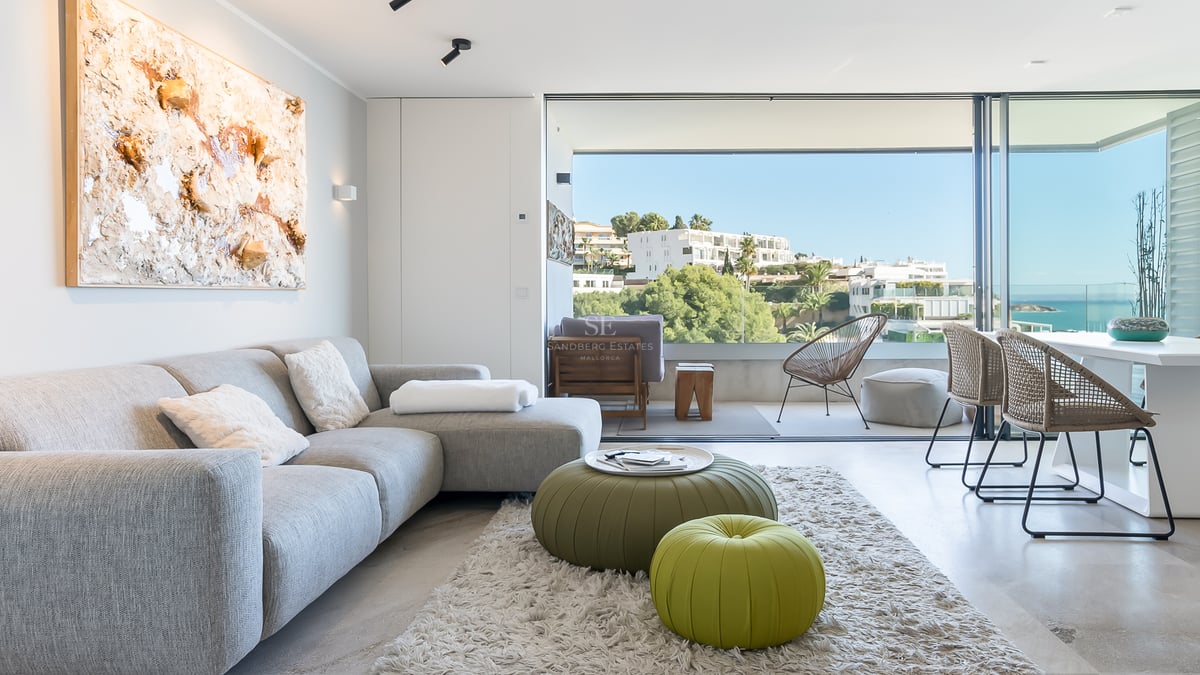 Helles, modernes Wohnzimmer mit grauem Sofa und grünen Sitzpoufs, bodentiefen Fenstern und Blick auf das Meer.