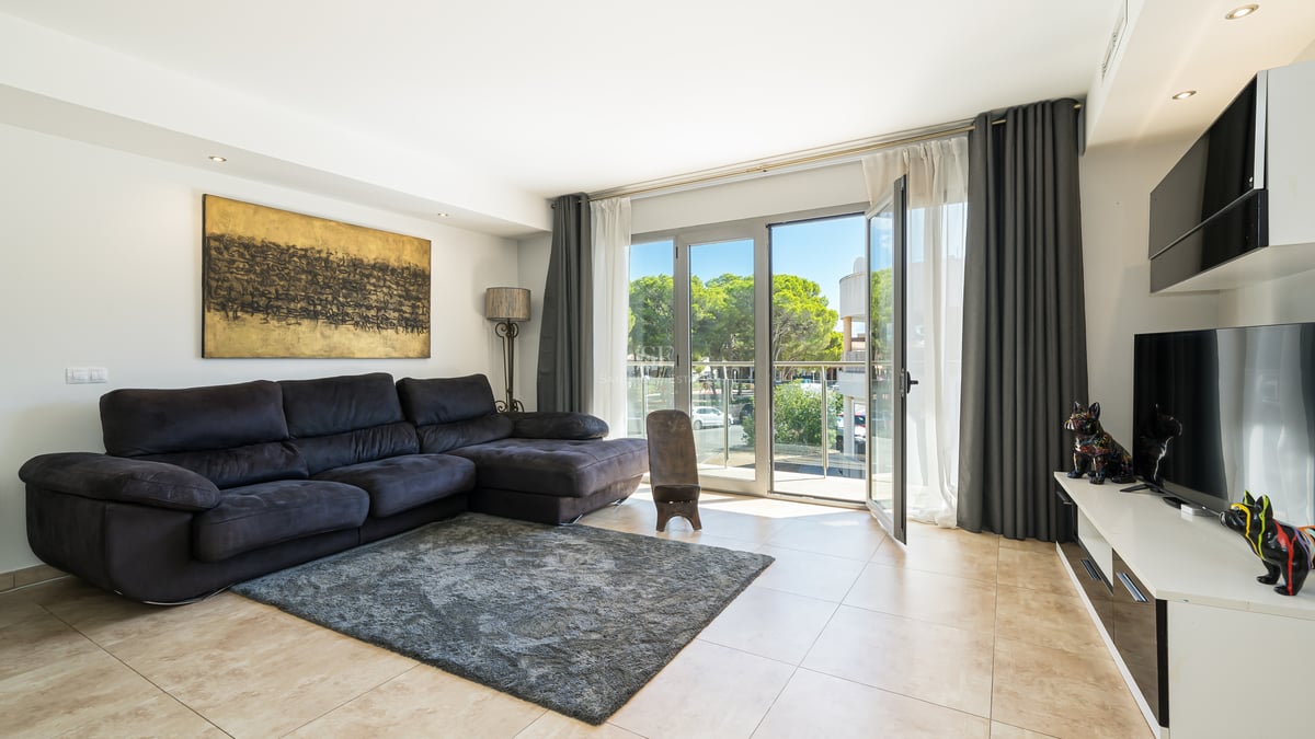 Ljust vardagsrum med en stor mörk soffa, grå matta och stora fönsterpartier som leder ut till en balkong.