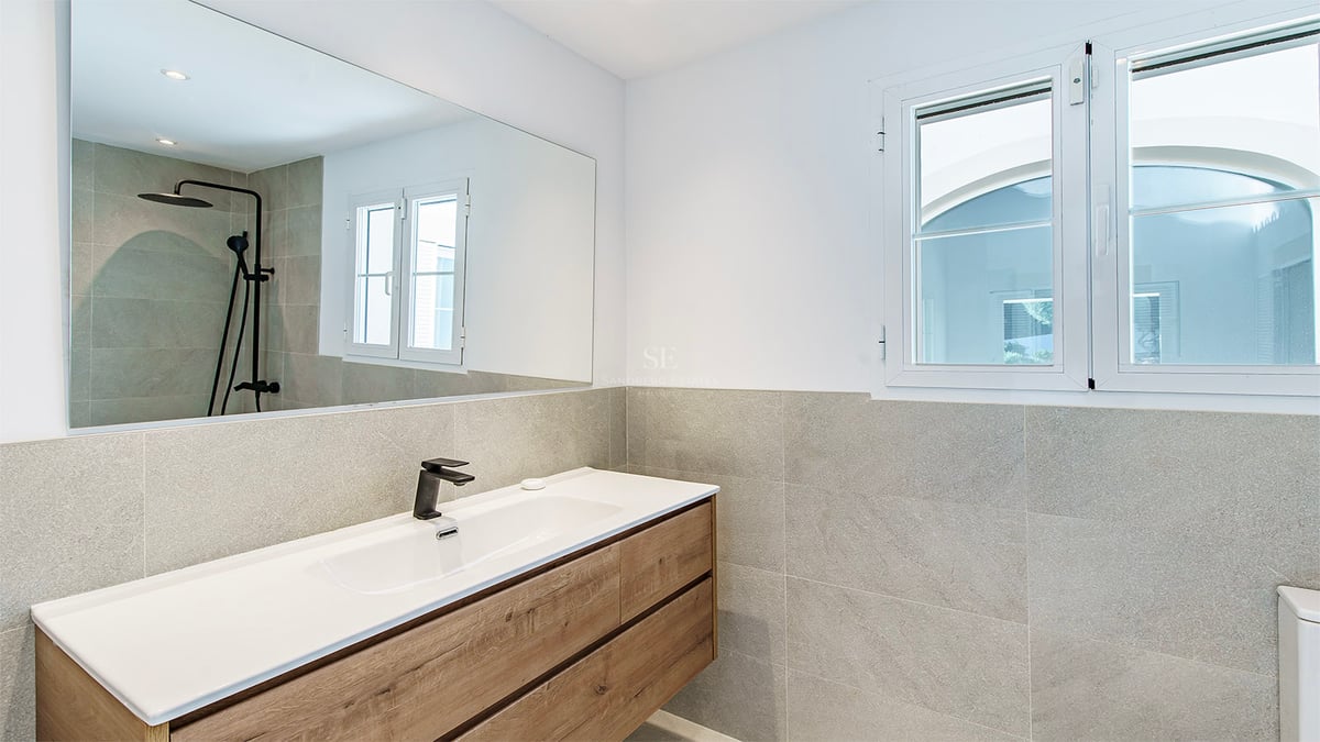 Modern badrumsinredning med träkommod, vitt tvättställ, svarta kranar och grått kakel.