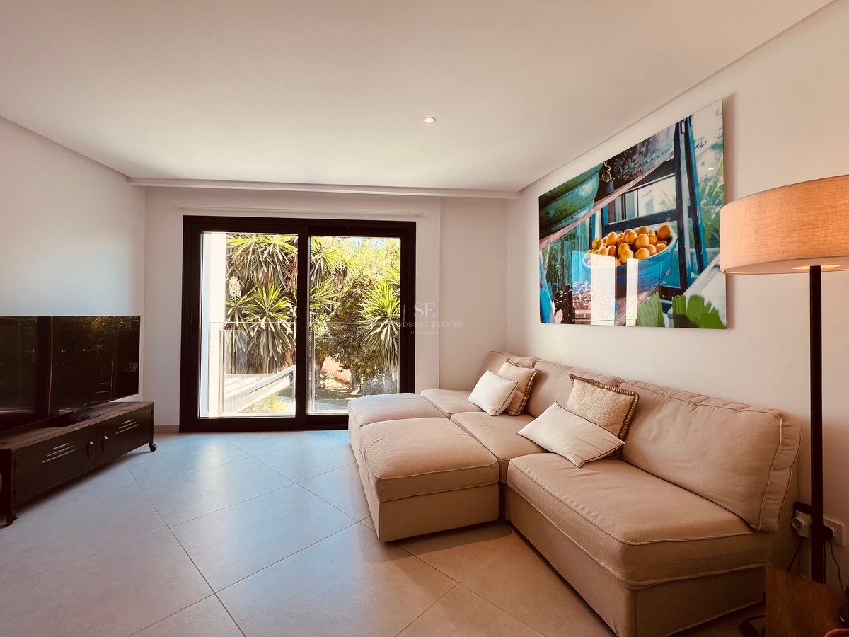 Salon moderne avec canapé d'angle beige, œuvre d'art murale et portes coulissantes donnant sur une terrasse arborée.