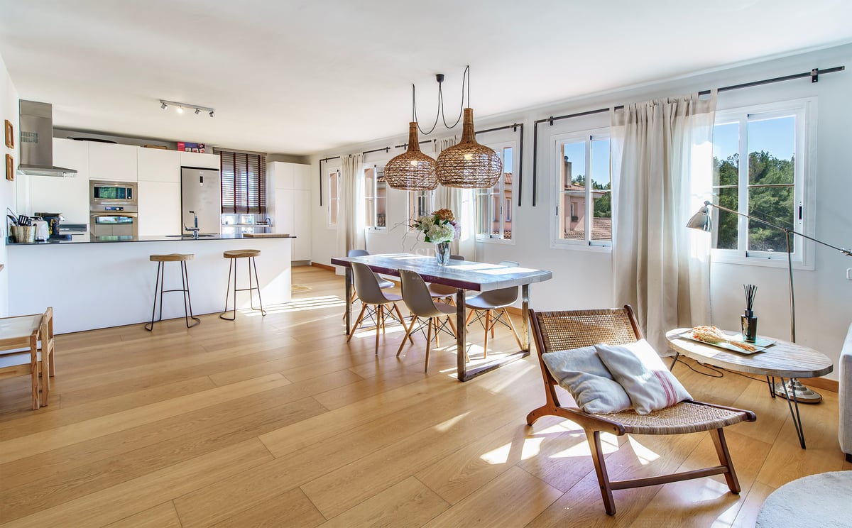 Soligt rum med öppen planlösning, vitt kök, matbord i trä och korglampor över ekparkett.