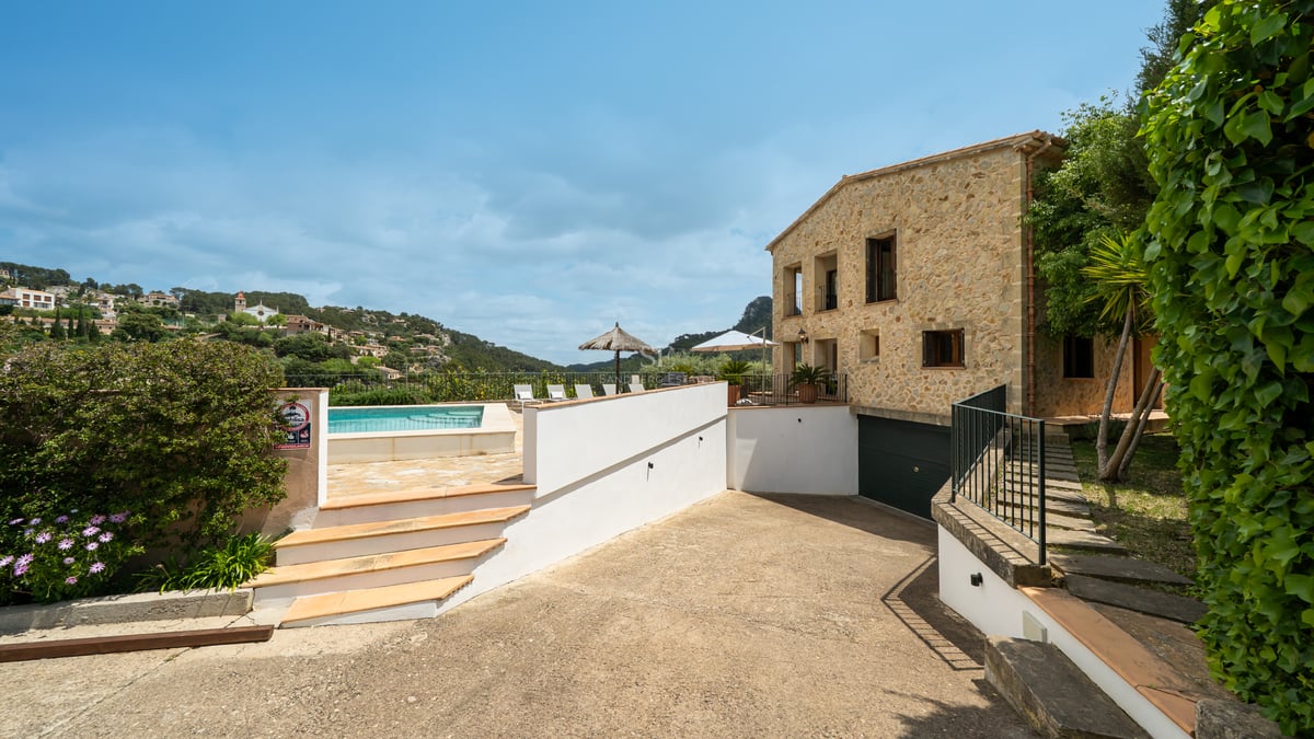Vue extérieure d'une villa en pierre avec piscine, terrasse ensoleillée et vue sur la montagne par temps clair.