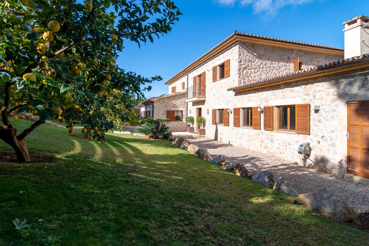 Exteriör av en naturstensvilla med träslutare, grön gräsmatta och citronträd under en klarblå himmel.