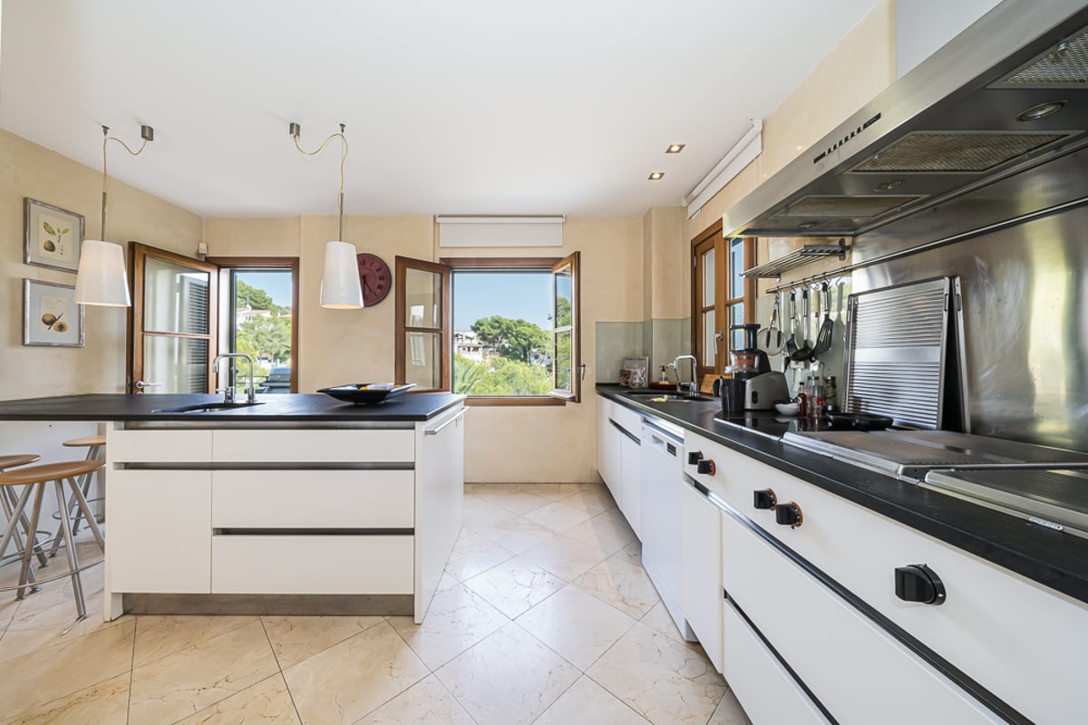 Modernt vitt kök med mörka bänkskivor, klinkergolv och fönster med utsikt över grönska.