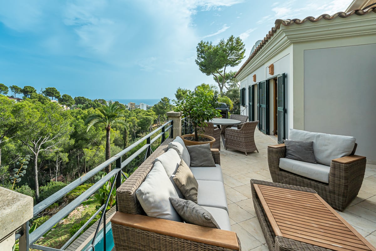 Rymlig terrass med loungemöbler i rotting och stengolv med utsikt över en grönskande dal och havet.