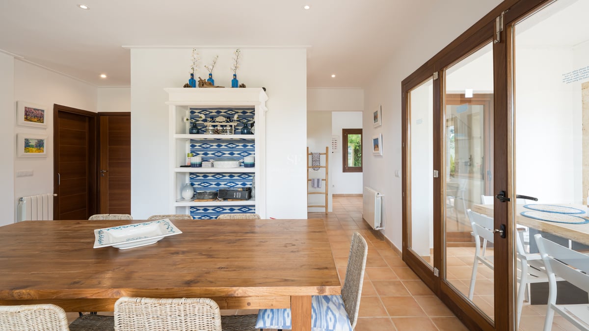Table à manger en bois massif et chaises en osier dans un intérieur lumineux avec sol en terre cuite et murs blancs.