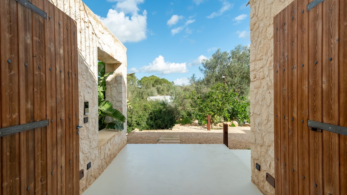 Entré med ljusa naturstensväggar som leder till en lummig medelhavsträdgård under en blå himmel.