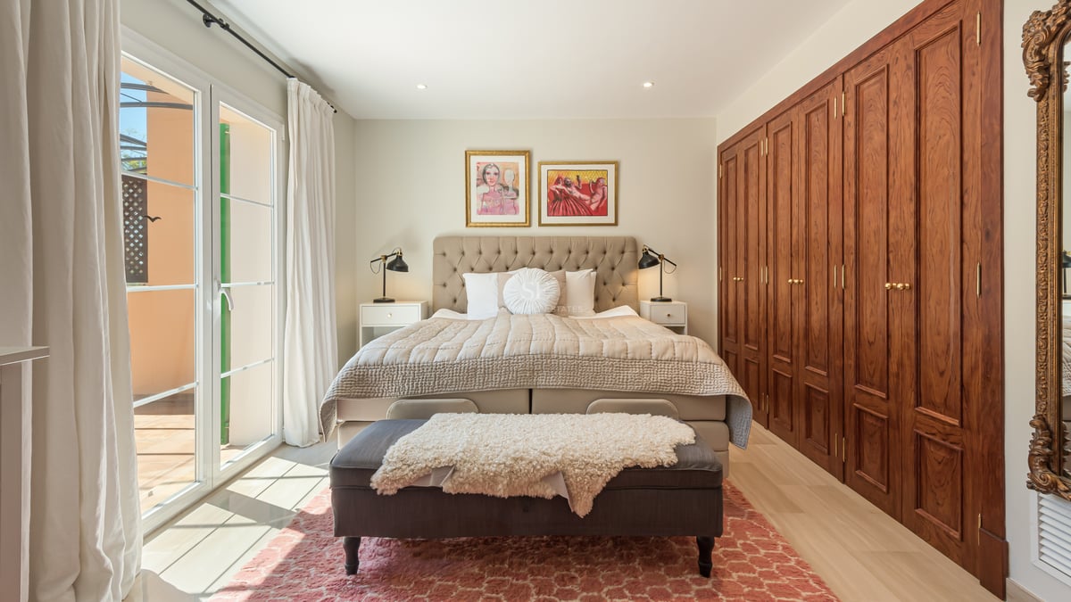 Sun-drenched master bedroom with tufted headboard, large wooden wardrobes, and glass doors opening to a terrace.