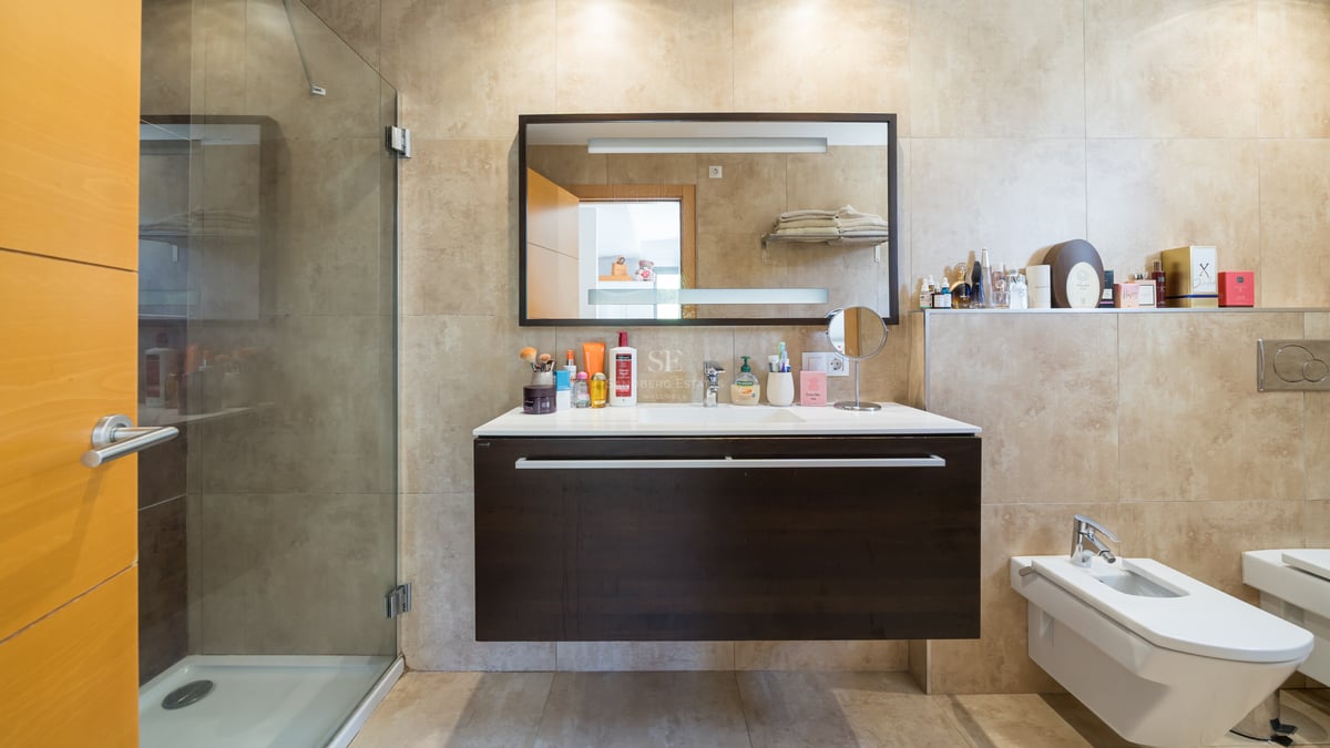 Modernt badrum med glasdusch, mörk svävande kommod och beige kakel.