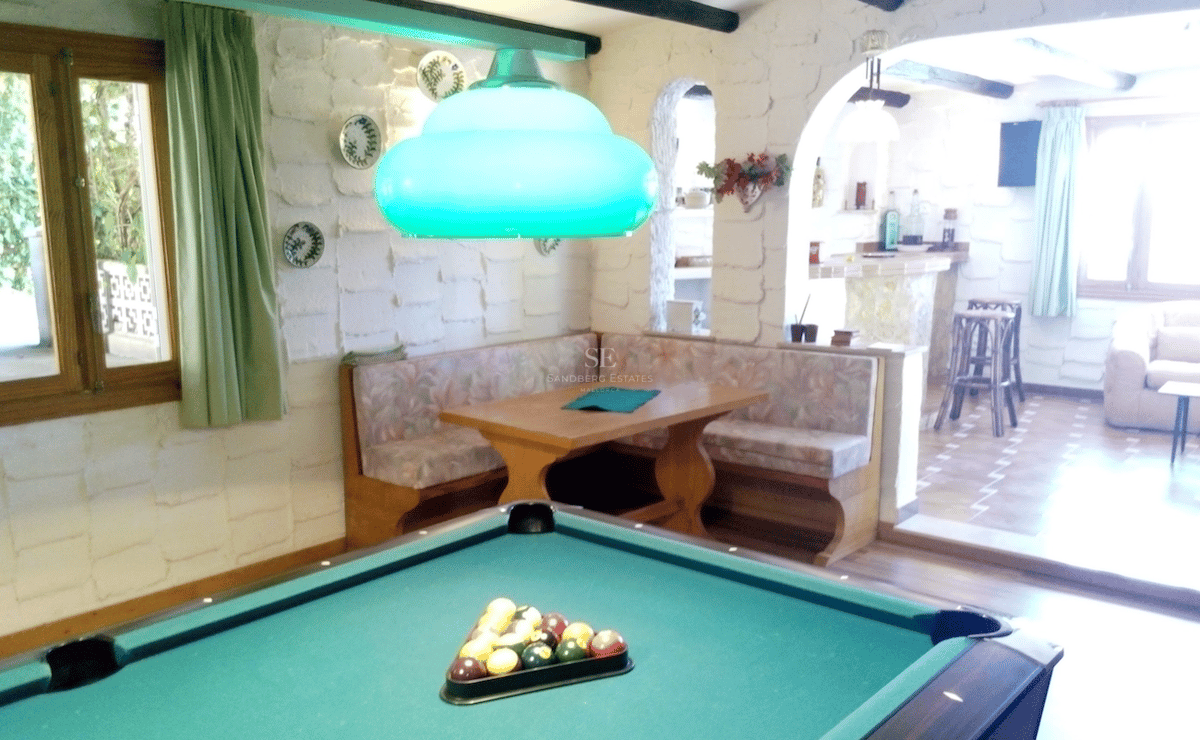 Sala de juegos rústica con mesa de billar, rincón de asientos de madera y paredes de piedra blanca.