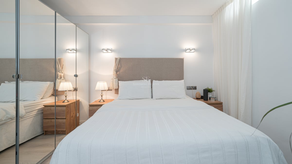 A bright modern bedroom featuring a beige upholstered headboard, white linens, and large floor-to-ceiling mirrored closets.