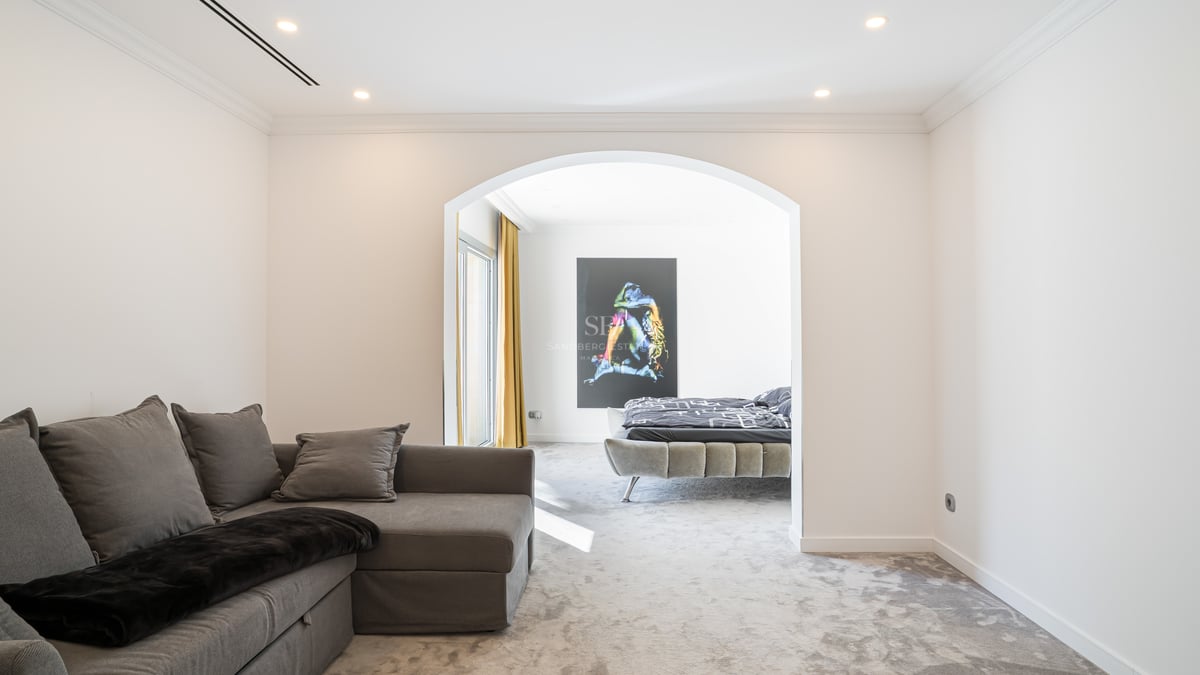 Modernes Schlafzimmer durch einen Rundbogen gesehen, mit grauem Polsterbett, weichem Teppich und großem Wandbild.