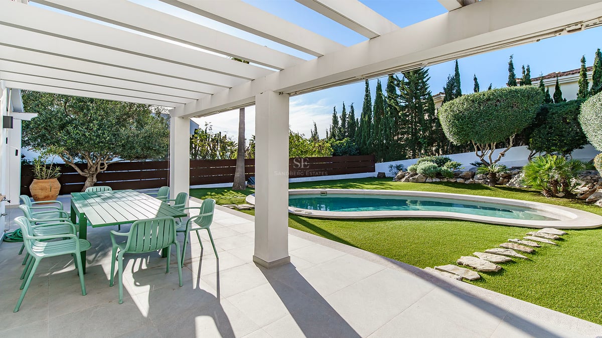 Skuggig terrass med grön matgrupp som vetter mot en pool och anlagd trädgård med cypresser.