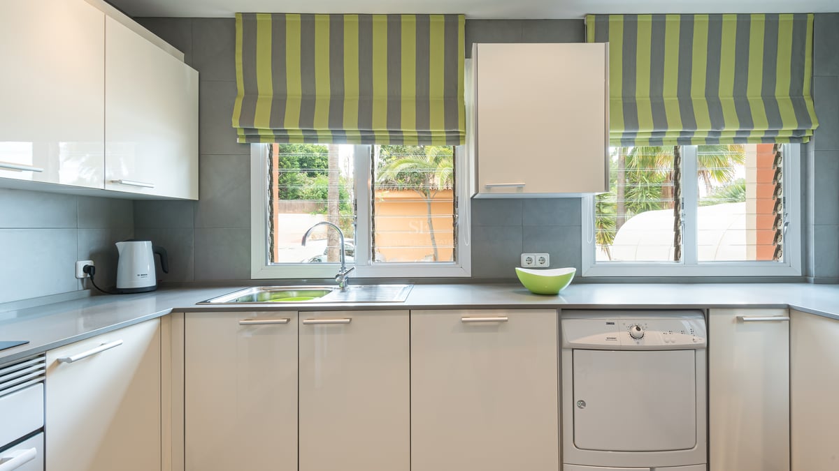 Contemporary kitchen featuring white glossy cabinets, grey countertops, and windows overlooking tropical palm trees.