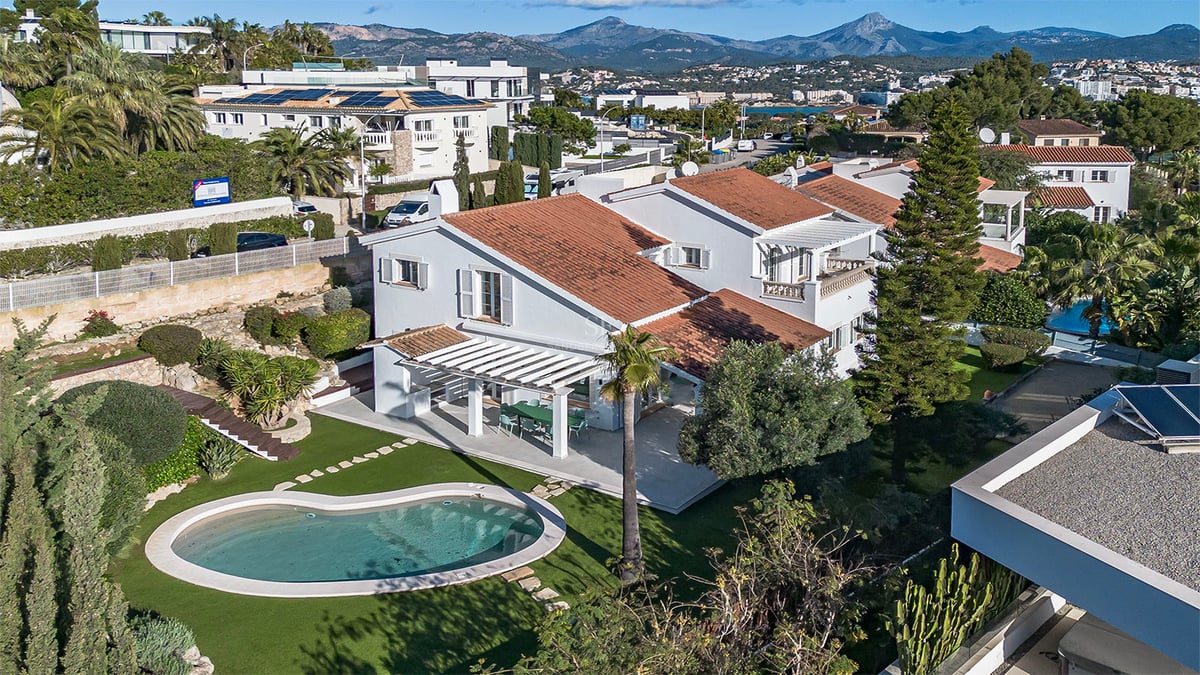 Flygfoto över en vit villa med tegeltak, njurformad pool och grön trädgård mot en bakgrund av berg.
