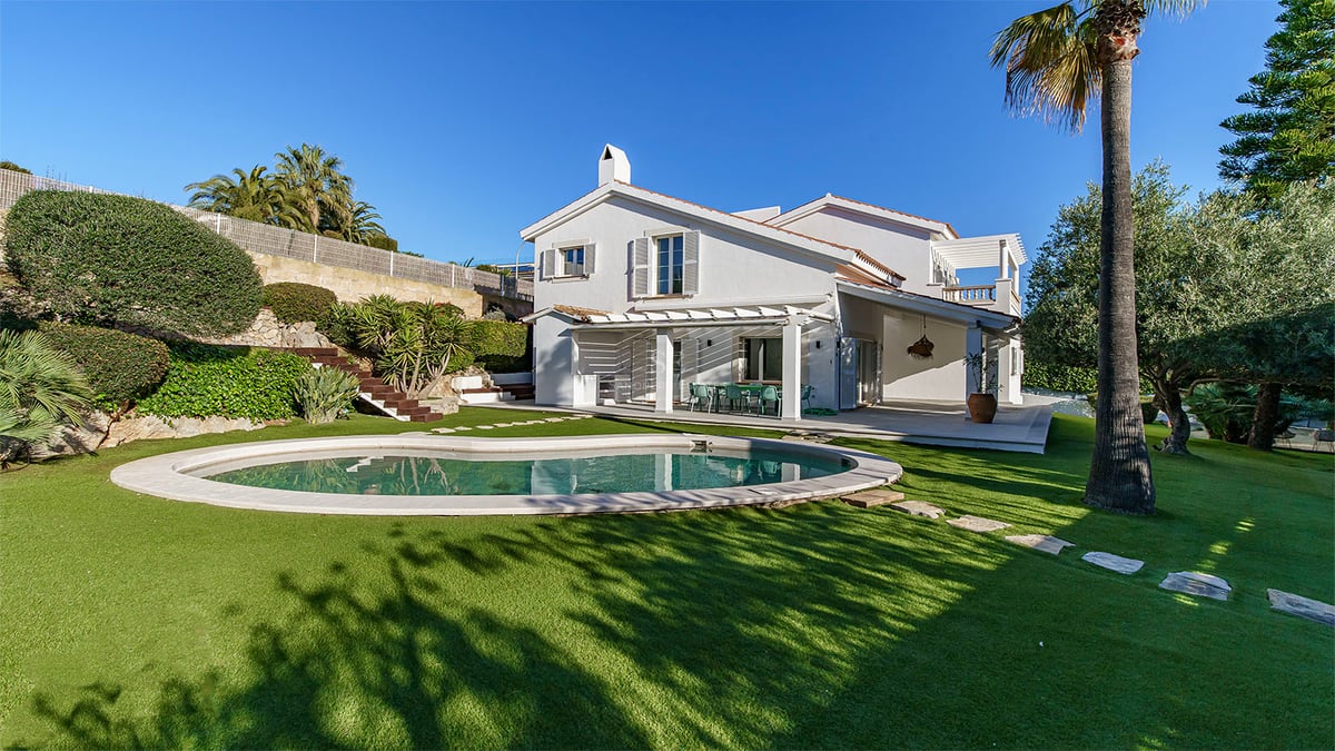 Vit tvåvåningsvilla med njurformad pool, grön gräsmatta och palmer under en klarblå himmel.