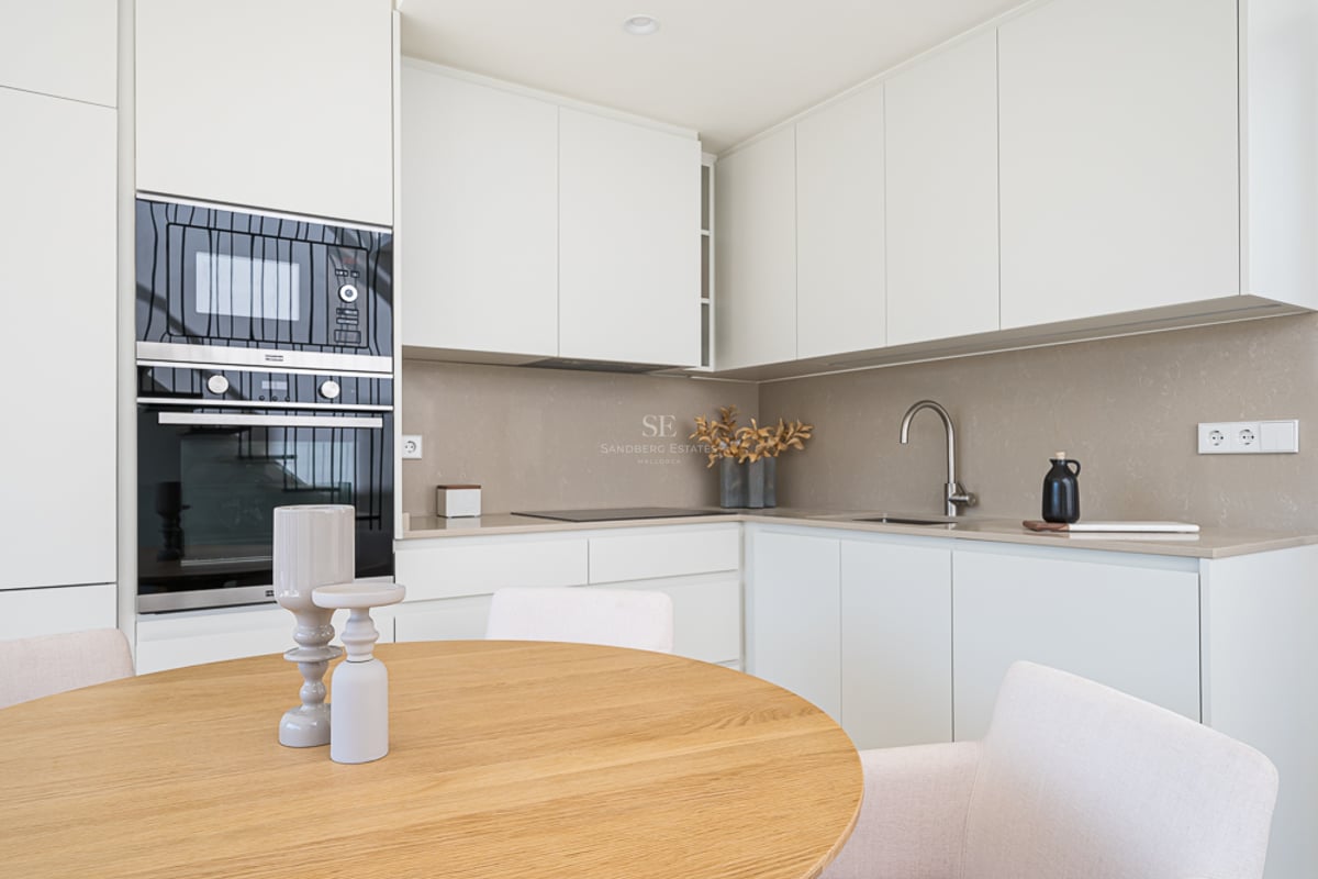Clean white kitchen cabinets with taupe backsplash, integrated ovens, and a round oak dining table in the foreground.