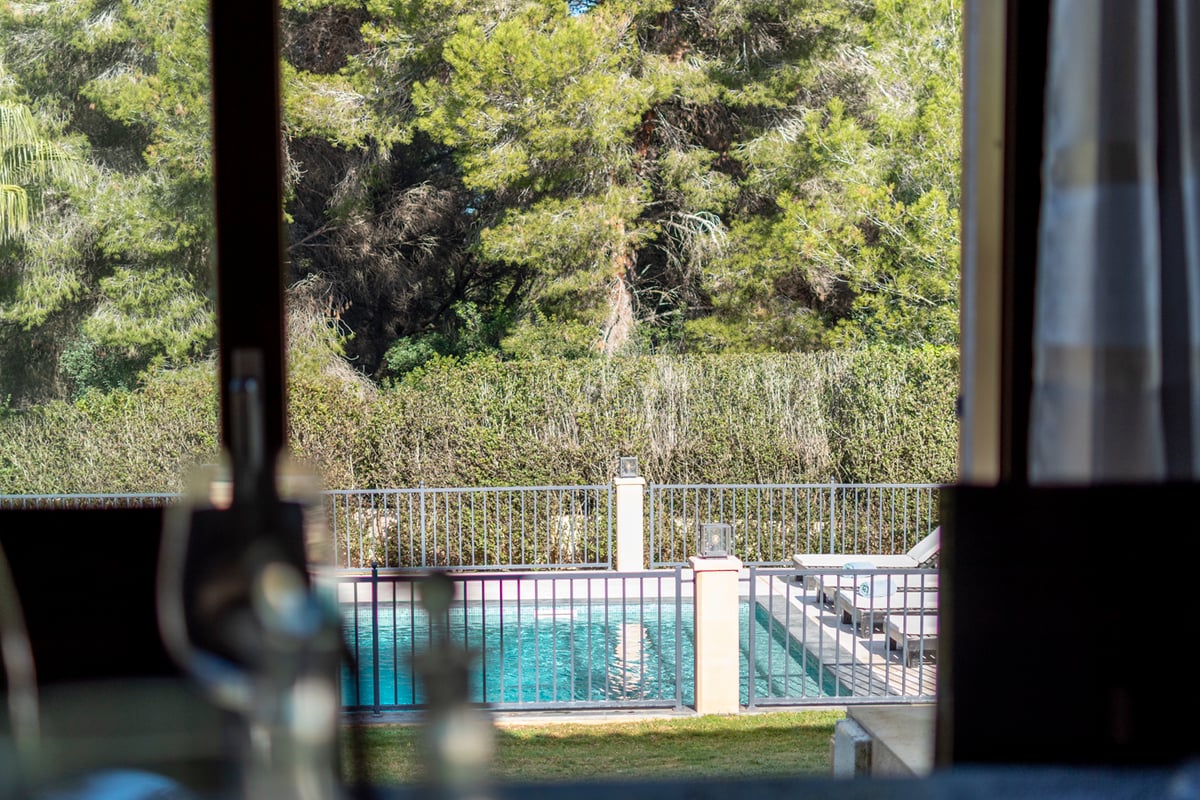 Utsikt genom ett fönster mot en inhägnad turkos pool omgiven av solstolar och höga tallar.