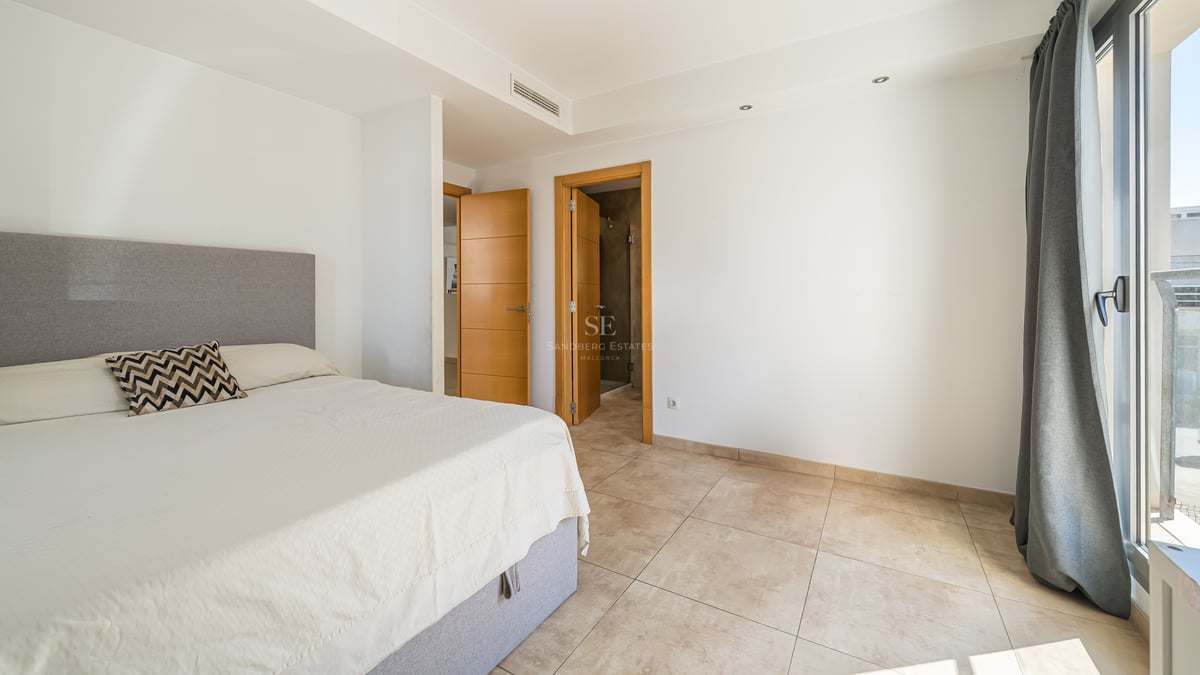 Ett soligt sovrum med grå säng, klinkergolv, vita väggar och trädörrar som leder till ett badrum.