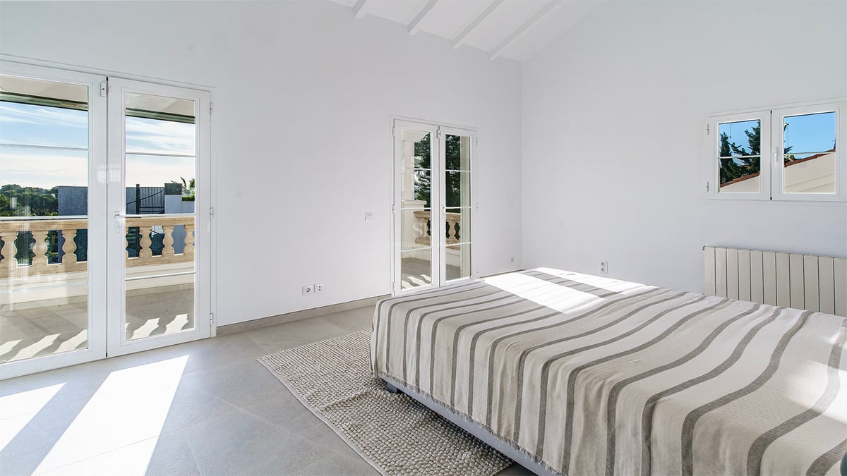 Minimalistiskt sovrum med randig säng, grått stengolv, högt tak med vita bjälkar och solig balkong.