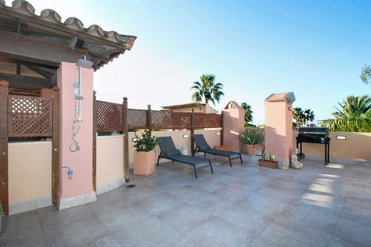 Stor medelhavsterrass med solstolar, utedusch, grill och ljusa stengolv under en klarblå himmel.