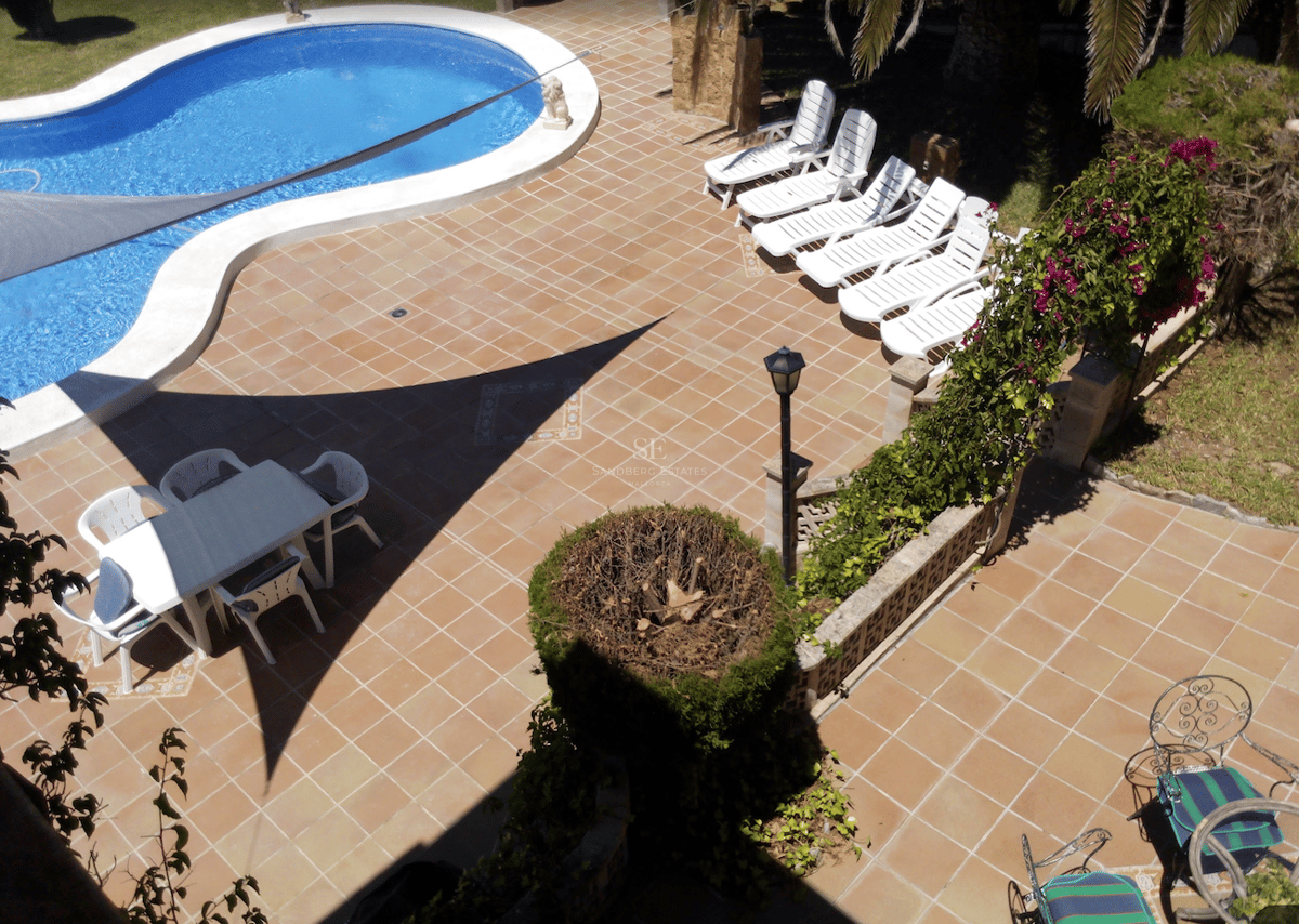 Vista aérea de una piscina azul en forma de riñón rodeada de baldosas de terracota, tumbonas blancas y zona de comedor.