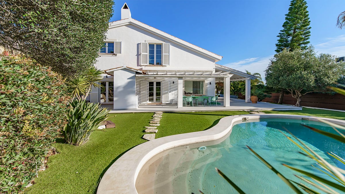 Baksida av en vit villa med en turkos pool, grön gräsmatta och en stenstig under en klarblå himmel.
