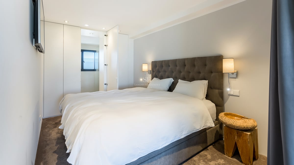 Modernes Schlafzimmer mit grauem gestepptem Kopfteil, weißen Einbauschränken und einem handgeschnitzten Holztisch.