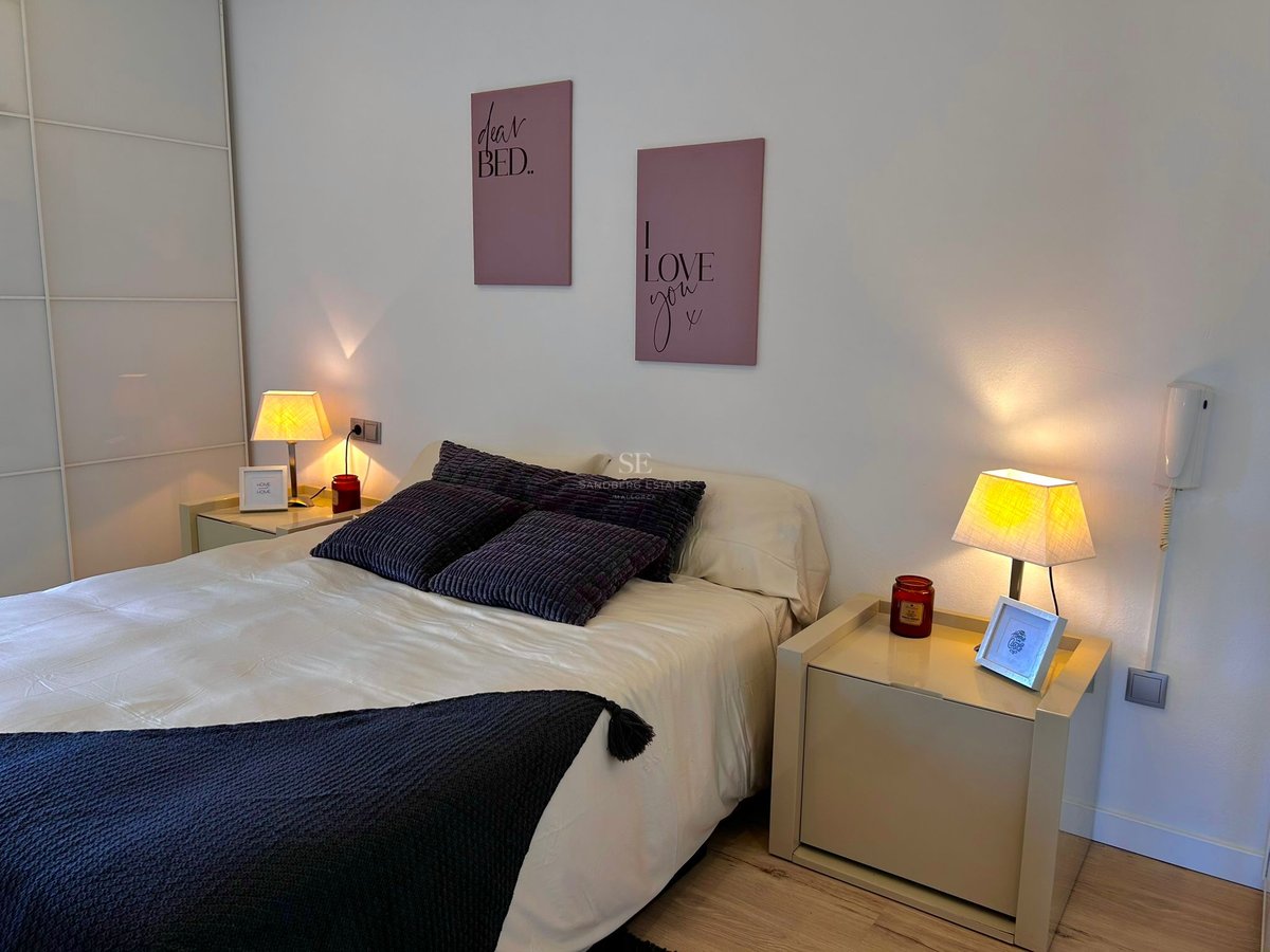 A cozy bedroom featuring a white bed with dark pillows, two bedside lamps glowing warmly, and pink wall art.