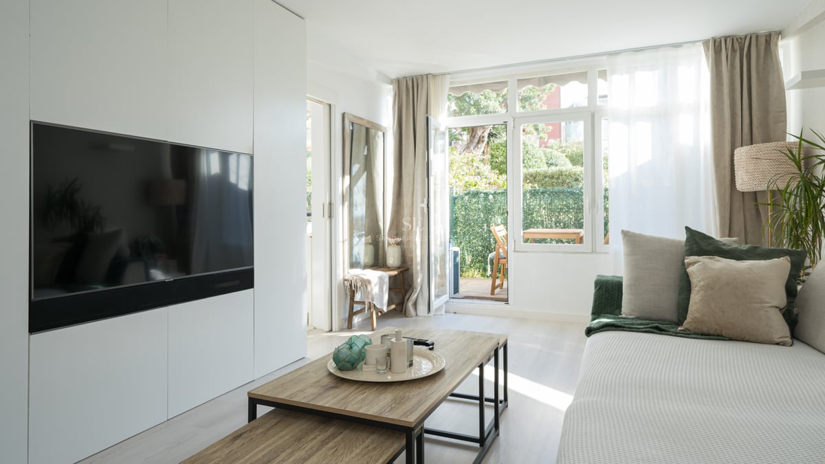 Modern living room with white wall unit, integrated TV, wooden coffee table, and glass door opening to a private garden.