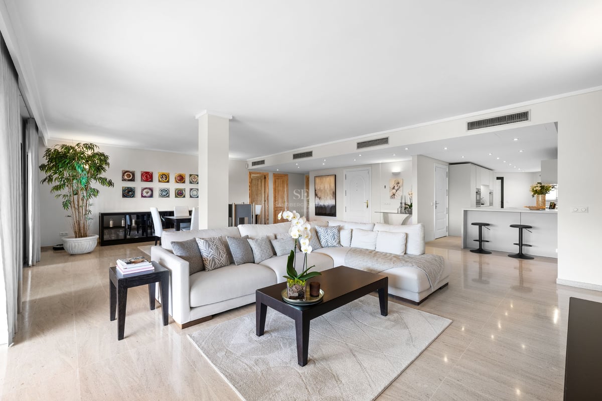 Spacieux salon avec canapé en L blanc, table basse en bois sombre, sol en marbre et cuisine intégrée.